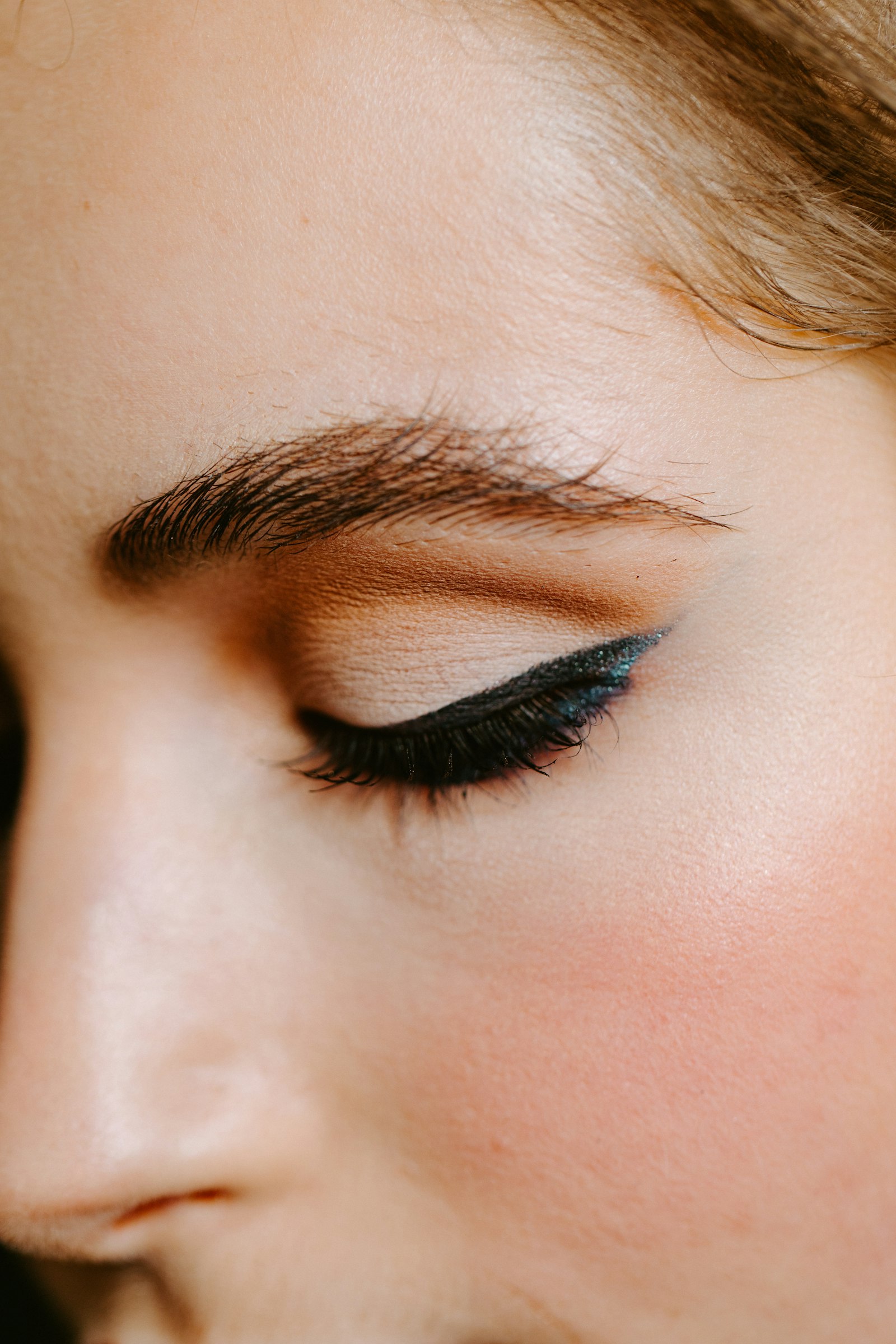 Close-up of a fluffy brushed-up eyebrow above a closed eye wearing teal glitter winged liner