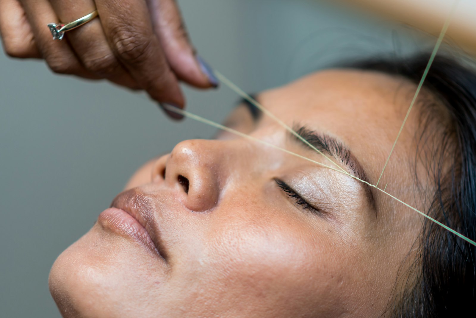 Brow threading in progress: a hand pulls cotton thread taut across the brow of a reclined client